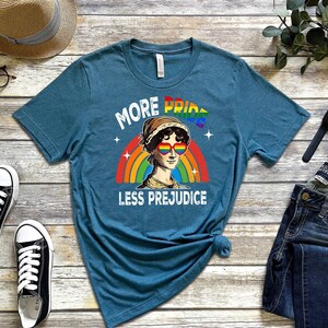 May include: Teal blue t-shirt featuring a rainbow and a cartoon image of a woman wearing rainbow sunglasses. The text on the shirt reads "More Pride Less Prejudice".
