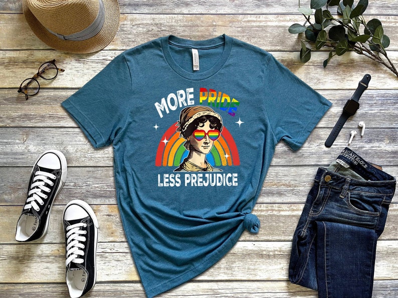 May include: Teal blue t-shirt featuring a rainbow and a cartoon image of a woman wearing rainbow sunglasses. The text on the shirt reads "More Pride Less Prejudice".
