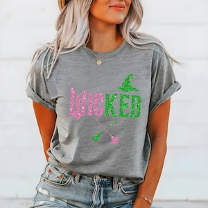 May include: A gray t-shirt with the word "WICKED" in pink and green glitter lettering. The design includes a green witch hat and crossed witch brooms. The shirt is a casual style.