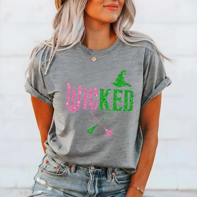 May include: A gray t-shirt with the word "WICKED" in pink and green glitter lettering. The design includes a green witch hat and crossed witch brooms. The shirt is a casual style.