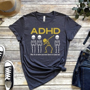 May include: Dark gray t-shirt with the word "ADHD" in gold letters and a skeleton graphic. The shirt also has the text "Why fit in when you were born to stand out?"