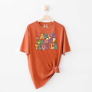 Puede incluir: Una camiseta naranja quemada con un estampado gr&aacute;fico que dice "Tacos and Tequila" en letras coloridas de estilo retro. El gr&aacute;fico tambi&eacute;n incluye ilustraciones de tacos, una margarita y una lima.