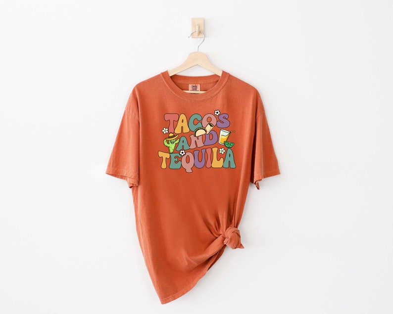 Puede incluir: Una camiseta naranja quemada con un estampado gr&aacute;fico que dice "Tacos and Tequila" en letras coloridas de estilo retro. El gr&aacute;fico tambi&eacute;n incluye ilustraciones de tacos, una margarita y una lima.