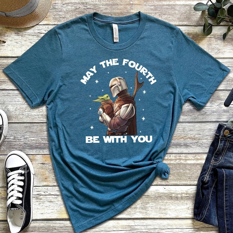 May include: A teal blue t-shirt with a white graphic of a Mandalorian character holding a baby Yoda. The text "May the Fourth Be With You" is printed in white on the shirt.