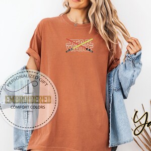 May include: A rust-coloured t-shirt with the words "STAR WARS" embroidered in white. Two crossed lightsabers are embroidered above the text in red and yellow.