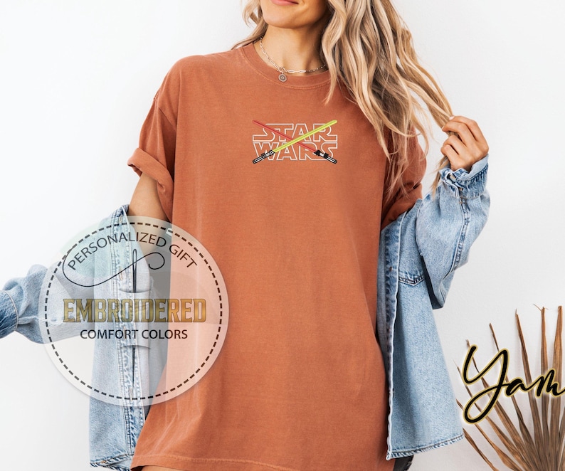 May include: A rust-coloured t-shirt with the words "STAR WARS" embroidered in white. Two crossed lightsabers are embroidered above the text in red and yellow.