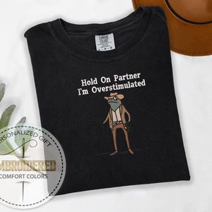 May include: Black Comfort Colors t-shirt with the embroidered phrase "Hold On Partner I'm Overstimulated" and a cartoon frog wearing a cowboy hat and vest. The shirt has a crew neck and short sleeves. The shirt is a personalized gift.