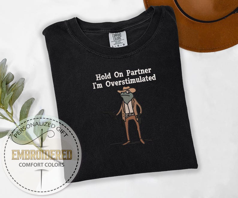May include: Black Comfort Colors t-shirt with the embroidered phrase "Hold On Partner I'm Overstimulated" and a cartoon frog wearing a cowboy hat and vest. The shirt has a crew neck and short sleeves. The shirt is a personalized gift.