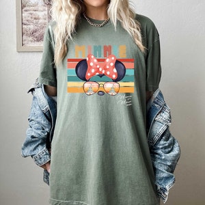 Op de afbeelding: Een groen T-shirt met een retro-stijl afbeelding van Minnie Mouse die een zonnebril draagt met de tekst "Minnie Mouse 2023" eronder. De afbeelding heeft een regenboog van kleuren en een witte stippen strik.