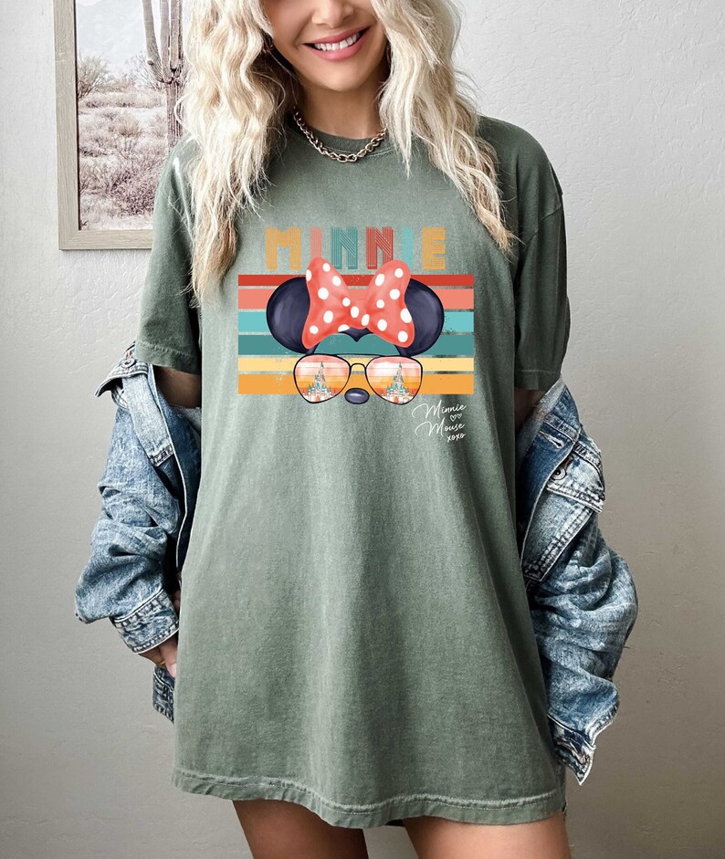 Op de afbeelding: Een groen T-shirt met een retro-stijl afbeelding van Minnie Mouse die een zonnebril draagt met de tekst "Minnie Mouse 2023" eronder. De afbeelding heeft een regenboog van kleuren en een witte stippen strik.