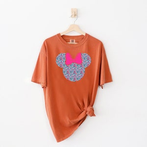 以下が含まれることがあります： さび色のTシャツに、花柄のミッキーマウスの頭のデザインがプリントされています。リボンはピンクです。