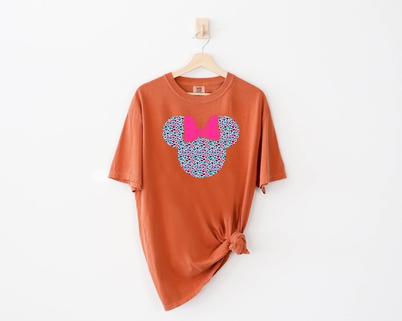 以下が含まれることがあります： さび色のTシャツに、花柄のミッキーマウスの頭のデザインがプリントされています。リボンはピンクです。