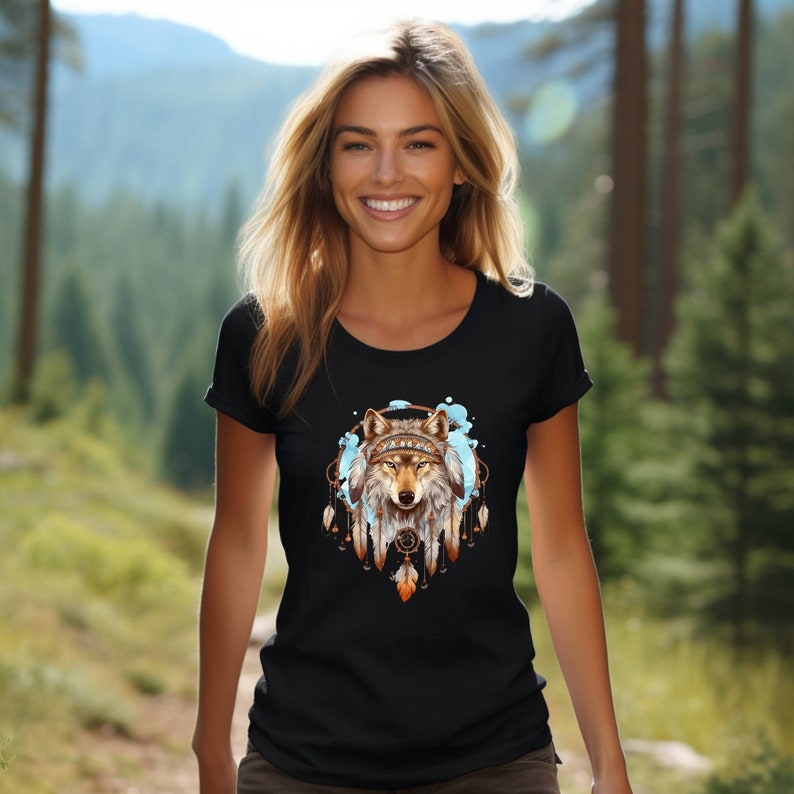 Pode incluir: Camiseta preta com a cabe&ccedil;a de um lobo dentro de um apanhador de sonhos com penas. O lobo est&aacute; usando uma faixa de contas. O apanhador de sonhos &eacute; cercado por respingos azuis como aquarela.