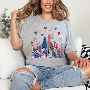 May include: A grey t-shirt with a patriotic castle design, fireworks, and heart-shaped details. The shirt features princesses in red, white, and blue dresses, ideal for a themed celebration or event.