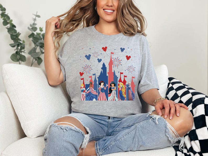 May include: A grey t-shirt with a patriotic castle design, fireworks, and heart-shaped details. The shirt features princesses in red, white, and blue dresses, ideal for a themed celebration or event.