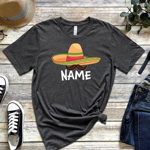 K&ouml;nnte beinhalten: Ein dunkelgraues T-Shirt mit einem Cartoon-Bild eines Sombreros, eines Schnurrbartes und dem Wort "NAME".