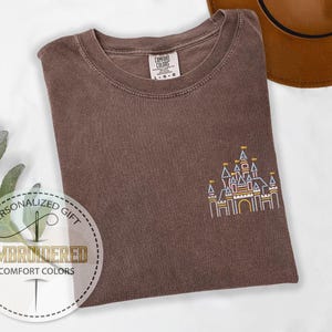 May include: A brown t-shirt with an embroidered outline of a castle in yellow, blue, and red. The text "Personalized Gift" and "Embroidered Comfort Colors" is on a white circle.