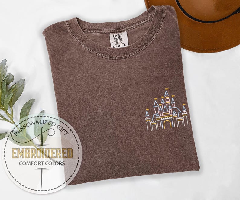 May include: A brown t-shirt with an embroidered outline of a castle in yellow, blue, and red. The text "Personalized Gift" and "Embroidered Comfort Colors" is on a white circle.