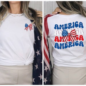 K&ouml;nnte beinhalten: Wei&szlig;es T-Shirt mit einer rot-wei&szlig;-blauen Schleife auf der Vorderseite und dem Wort "AMERICA" viermal in einer Retro-Schrift auf der R&uuml;ckseite.