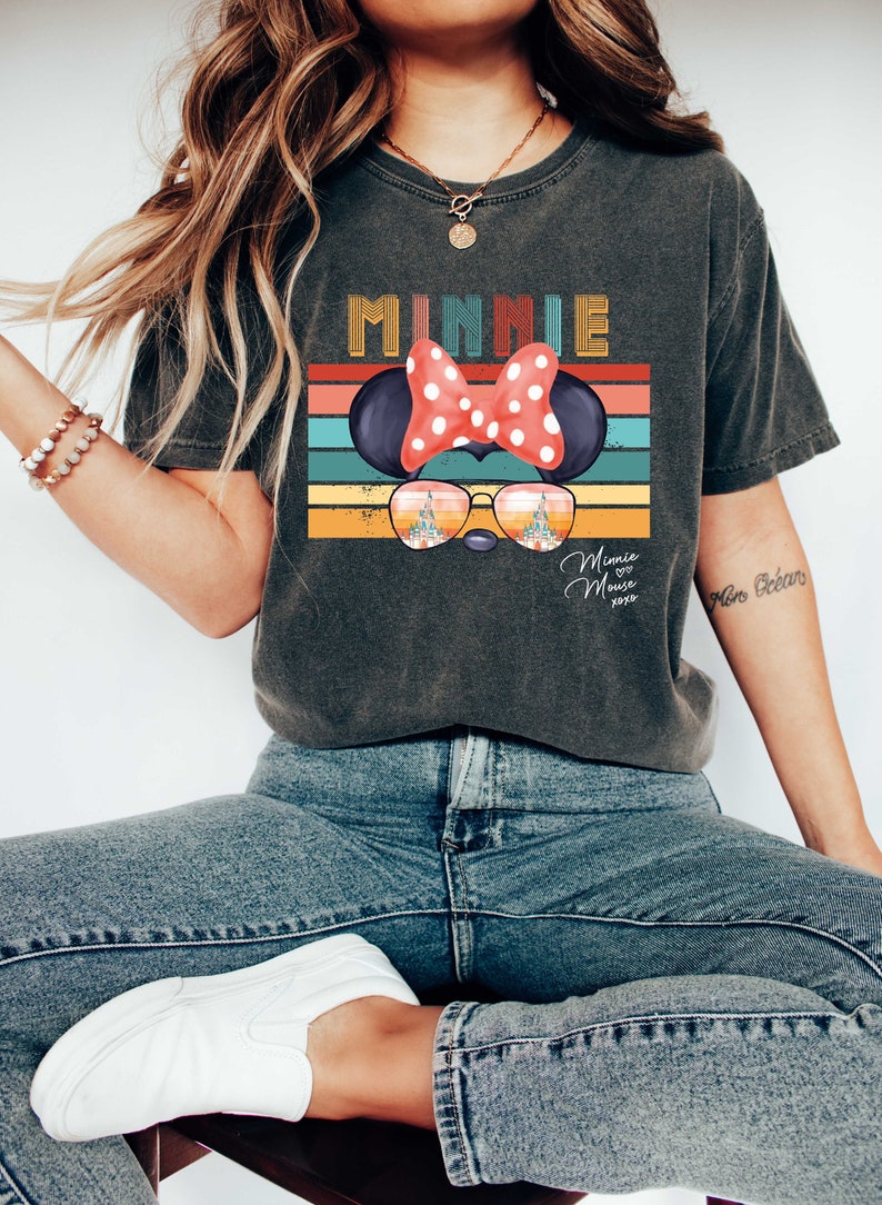 Op de afbeelding: Een donkergrijs T-shirt met een vintage-stijl grafische afbeelding van Minnie Mouse die een zonnebril draagt ​​met het kasteel van Assepoester weerspiegeld in de glazen. De afbeelding bevat ook de tekst "Minnie Mouse" en "xoxo".