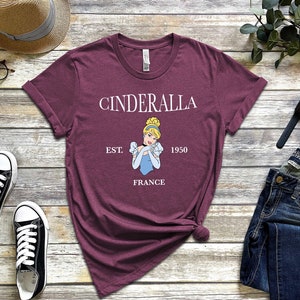 Peut inclure: T-shirt bordeaux avec un motif Cendrillon. Le design comprend le mot "Cinderalla", l'ann&eacute;e "1950" et le mot "France". Le t-shirt est pr&eacute;sent&eacute; avec des baskets noires, un chapeau de paille et un jean bleu.