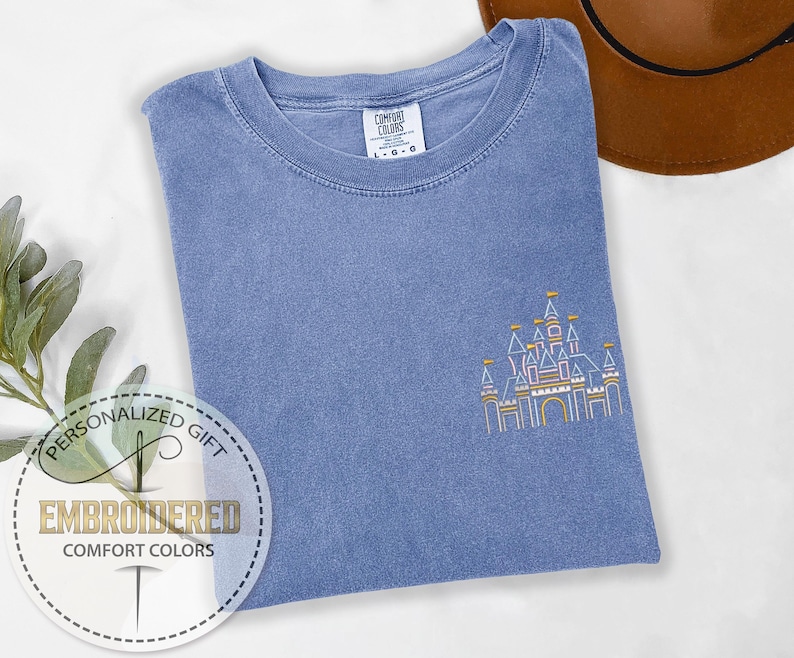 May include: A light blue t-shirt with an embroidered outline of a castle in pink, yellow, and white. The text "Personalized Gift" and "Embroidered Comfort Colors" is printed on a white circle.