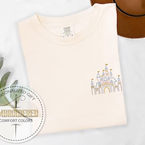 May include: A white t-shirt with an embroidered design of a pink, blue, and yellow castle. The text "PERSONALIZED GIFT" and "EMBROIDERED COMFORT COLORS" is on a white circle with a needle and thread design.