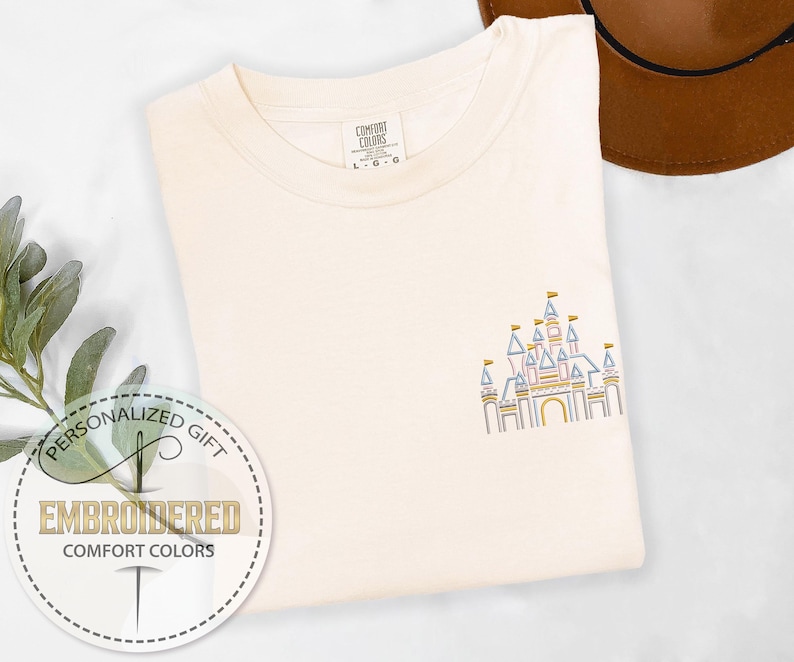 May include: A white t-shirt with an embroidered design of a pink, blue, and yellow castle. The text "PERSONALIZED GIFT" and "EMBROIDERED COMFORT COLORS" is on a white circle with a needle and thread design.