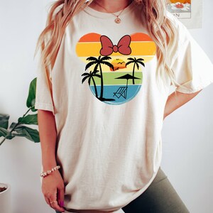 May include: A white t-shirt with a retro-style graphic of a Mickey Mouse head silhouette. The graphic features a sunset, palm trees, a beach chair, and a red bow.