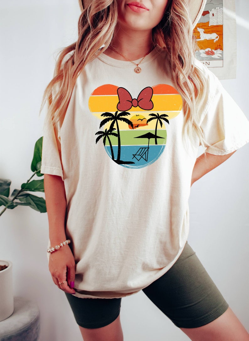 May include: A white t-shirt with a retro-style graphic of a Mickey Mouse head silhouette. The graphic features a sunset, palm trees, a beach chair, and a red bow.