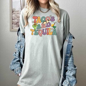 Puede incluir: Una camiseta verde claro con el texto "TACOS AND TEQUILA" en letras retro de colores. El texto est&aacute; rodeado de peque&ntilde;as ilustraciones de tacos, un cactus, una margarita y limas.