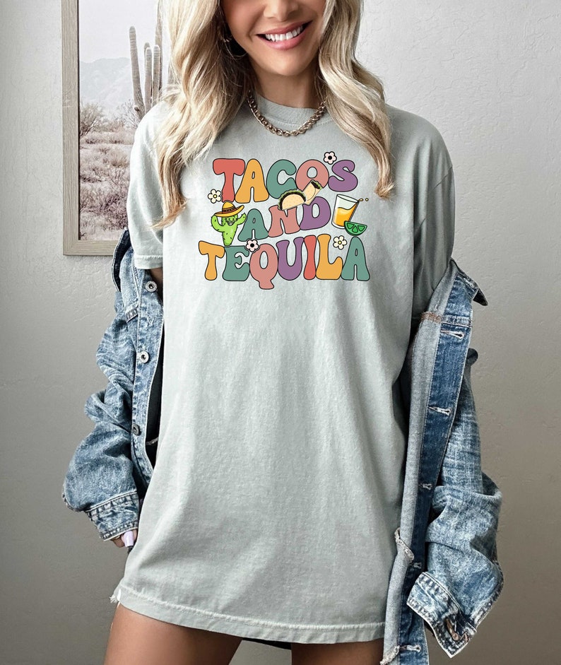 Puede incluir: Una camiseta verde claro con el texto "TACOS AND TEQUILA" en letras retro de colores. El texto est&aacute; rodeado de peque&ntilde;as ilustraciones de tacos, un cactus, una margarita y limas.