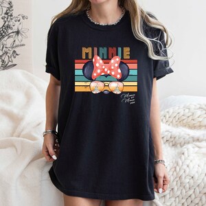 Op de afbeelding: Zwart T-shirt met een retro-stijl grafische afbeelding van Minnie Mouse met een zonnebril en een rode strik. De afbeelding bevat de tekst "Minnie" en "Mouse House" in een vintage lettertype.