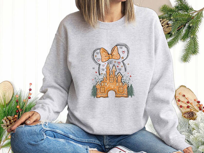 Puede incluir: Una sudadera gris con un castillo de jengibre y un lazo. El castillo est&aacute; decorado con chispas blancas y el lazo es rojo y blanco.