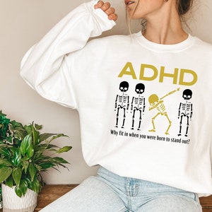 May include: White sweatshirt with the word "ADHD" in gold letters and a graphic of four skeletons. One skeleton is dabbing, and the text "Why fit in when you were born to stand out?" is below the skeletons.