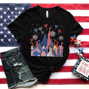 May include: Black t-shirt with a patriotic design featuring cartoon princesses, fireworks, and a castle silhouette. The shirt is displayed on an American flag, alongside denim shorts. A small sign reads "Home of the Brave."