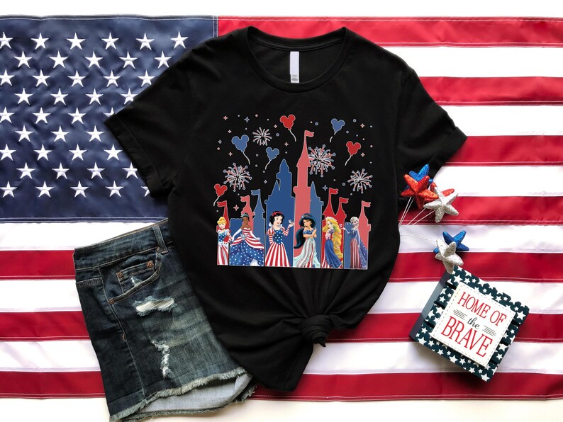 May include: Black t-shirt with a patriotic design featuring cartoon princesses, fireworks, and a castle silhouette. The shirt is displayed on an American flag, alongside denim shorts. A small sign reads "Home of the Brave."