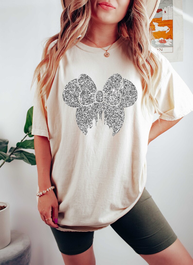 May include: A white oversized t-shirt with a silver glitter bow design. The shirt is worn with dark green biker shorts.