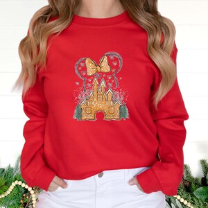 Puede incluir: Sudadera roja de manga larga con un gr&aacute;fico de un castillo de jengibre y un lazo, con un fondo nevado.