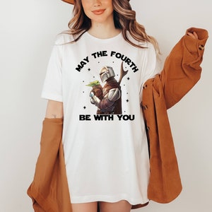 May include: White t-shirt with a black graphic of a Mandalorian character holding a baby Yoda with the text "May the Fourth Be With You".