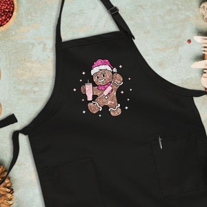Peut inclure: Tablier de cuisine noir avec un bonhomme en pain d'&eacute;pices tenant une boisson rose et portant un chapeau de P&egrave;re No&euml;l rose et blanc. Le bonhomme en pain d'&eacute;pices est entour&eacute; d'&eacute;toiles scintillantes roses.