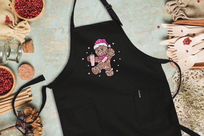 Peut inclure: Tablier de cuisine noir avec un bonhomme en pain d'&eacute;pices tenant une boisson rose et portant un chapeau de P&egrave;re No&euml;l rose et blanc. Le bonhomme en pain d'&eacute;pices est entour&eacute; d'&eacute;toiles scintillantes roses.