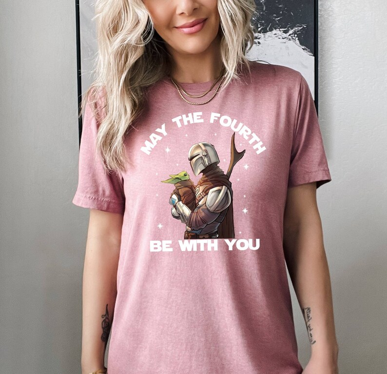 May include: A pink t-shirt with a white graphic of a Mandalorian character holding a baby Yoda. The text "May the Fourth Be With You" is printed on the shirt.