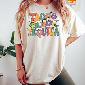 Puede incluir: Una camiseta blanca con el texto "Tacos and Tequila" en una fuente retro colorida. El texto est&aacute; rodeado de ilustraciones de tacos, un cactus y una margarita.