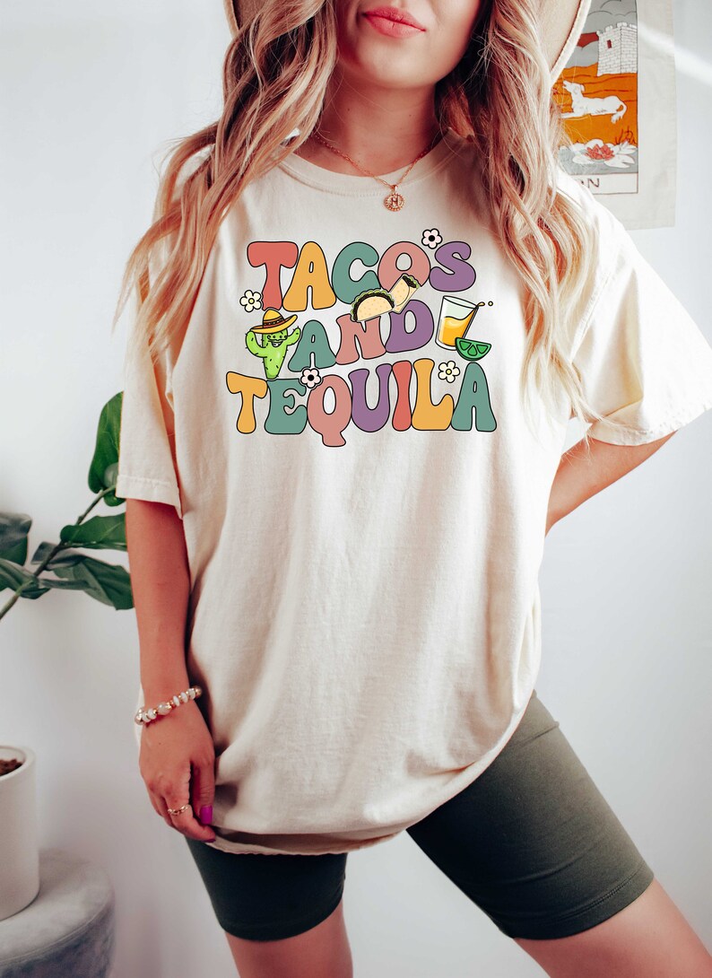 Puede incluir: Una camiseta blanca con el texto "Tacos and Tequila" en una fuente retro colorida. El texto est&aacute; rodeado de ilustraciones de tacos, un cactus y una margarita.
