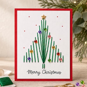 Puede incluir: Una tarjeta de felicitación blanca con borde rojo que presenta un diseño de árbol de Navidad bordado con hilo verde. El árbol está decorado con pequeñas cuentas de colores y una estrella dorada en la punta. El texto Merry Christmas está impreso en negro.