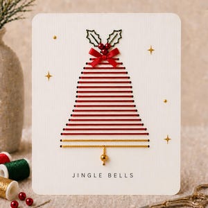Peut inclure: Une carte de vœux blanche ornée d'un motif de cloche réalisé avec du fil rouge et doré horizontal. La cloche est surmontée d'un nœud en ruban rouge, de feuilles de houx brodées vertes et de perles rouges. Des étoiles dorées et le texte JINGLE BELLS.