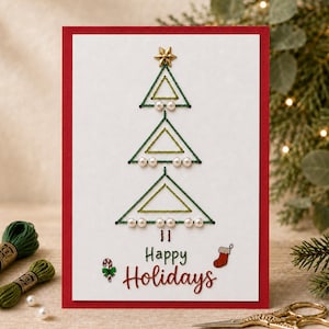 Peut inclure: Une carte de vœux blanche à bordure rouge ornée d'un sapin de Noël brodé en fil vert, agrémenté de perles blanches et d'une étoile dorée. Le texte Happy Holidays est imprimé en vert et rouge, accompagné d'une canne en sucre et d'une chaussette.