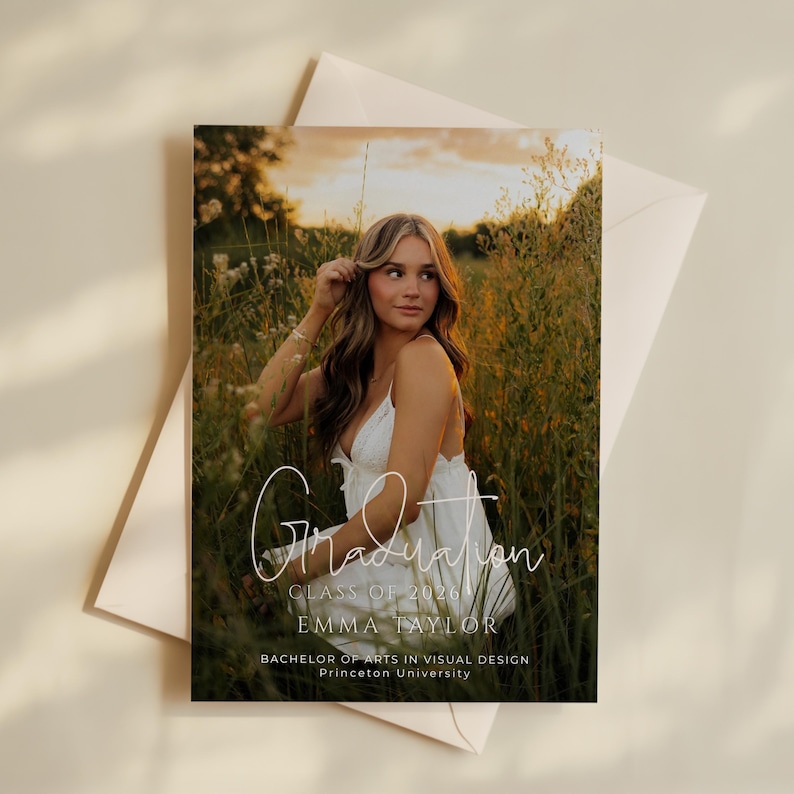 May include: Graduation announcement featuring a young woman in a white dress, seated in a field of tall grass. The card includes the word "Graduation" in script, the year "2026", the name "Emma Taylor", and her degree and university.