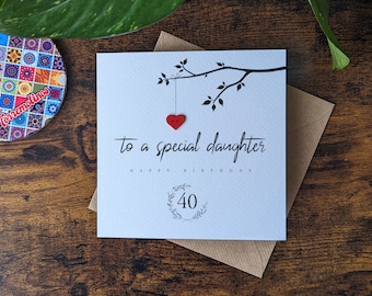 Puede incluir: Una tarjeta blanca con texto negro que dice "To a special daughter Happy Birthday 40". Un botón de corazón rojo cuelga de una rama negra dibujada en la tarjeta.